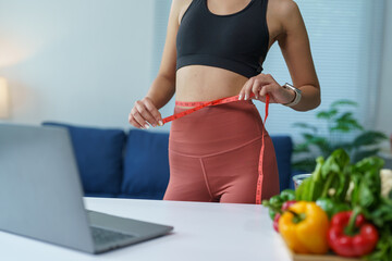 Young woman in sportswear measuring her waist with a tape measure while following an online diet...