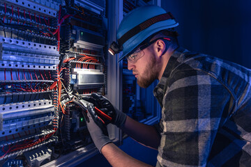 A male electrician works in a switchboard in overalls against the backdrop of emergency lighting.