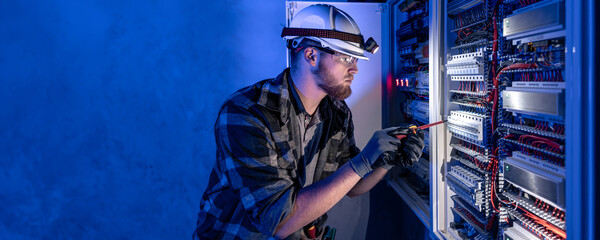 A male electrician works in a switchboard in overalls against the backdrop of emergency lighting.