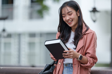 Smiling young asian woman enjoying her studies while reading a book outdoors, embracing a casual and relaxed atmosphere on a university campus during spring