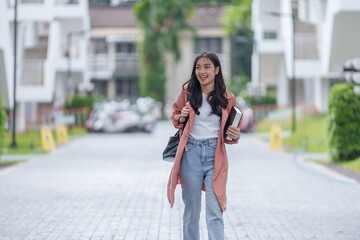 Fototapeta premium Cheerful young asian woman, dressed in casual attire, strides confidently through a university campus, books in hand, embodying the spirit of academic pursuit and youthful enthusiasm