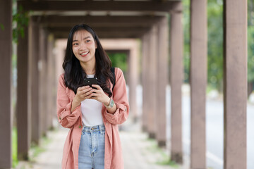 Happy university student using mobile phone app for texting, online shopping, social media, or playing mobile game while walking in covered walkway
