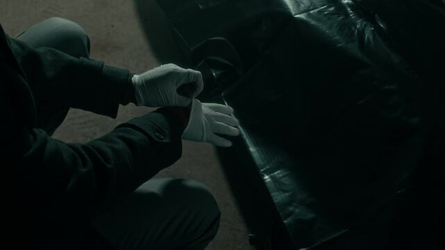 High angle cropped shot of Black male forensic expert putting on sterile gloves to examine lying dead body covered with plastic bag in dark gloomy setting