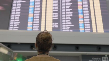 Woman traveler standing with back turned, scanning digital timetable screen at modern airport terminal, checking flight information and schedule - Powered by Adobe