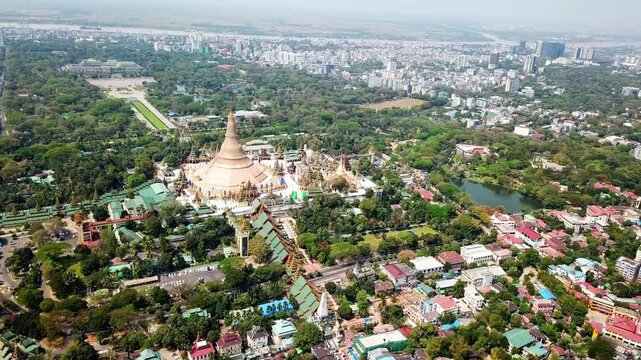 Breathtaking aerial view of the iconic Shwedagon Pagoda in Rangun, Myanmar, surrounded by lush greenery, urban structures, and tranquil lakes, showing blend of cultural heritage and modern city life
