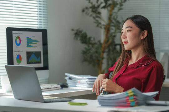 Young asian businesswoman relaxing and thinking at office desk after working hard analyzing financial data using laptop computer and examining charts and graphs on computer screen