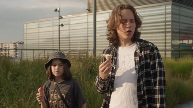 The older and younger brothers spend time together, walking and eating ice cream on a summer day. The boys walk slowly, talk amiably to each other and sit down to relax on a bench in the city park.