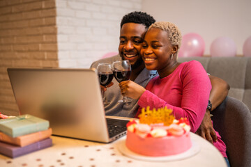Happy young couple celebrating woman's birthday at home. They are toasting with wine and having video call with family.