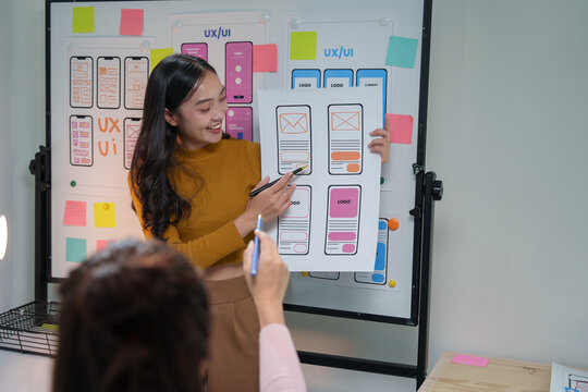 Asian web designer presenting a wireframe mockup of a mobile app interface during a collaborative meeting with her team in the office, fostering creativity and innovation