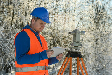 Surveyor using tablet and theodolite during winter land surveying in snowy landscape