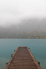 Obraz premium Small jetty towards a lake with fog-covered mountain range and turquoise water (South Island, New Zealand)