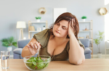 Portrait of unhappy sad fat woman sitting at table in kitchen in front of a bowl of green salad. Overweight girl eating healthy food at home. Nutrition, dieting and weight loss concept.