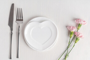 Top view of flat lay with heart shaped ceramic plate, pink carnation flowers and tableware of knife and fork served on white wooden table prepared for dinner for celebration of valentines day or date
