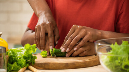 African American mother and little son preparing healthy meal, close up shot of mom helping son cut cucumber with knife, holding boy's hands.