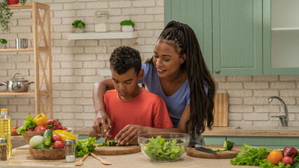 African American mother and little son preparing healthy meal, mom helping son cut cucumber with knife for salad, holding boy's hands.