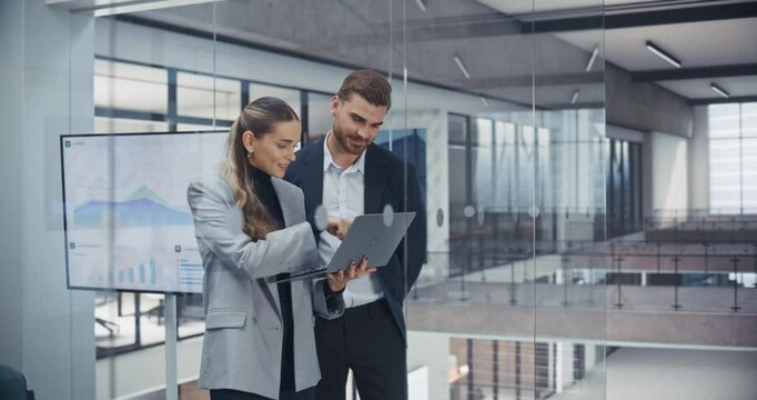 Two Young Businesspeople Standing in a Meeting Room, Inspecting Latest Financial Report on a Laptop Computer. Caucasian Male and Female Managers Discussing Next Steps to Benefit the Company