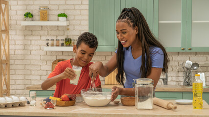 African American mother and little son preparing healthy meal, making dough for baking, mom mixing flour in bowl, boy holding glass of milk.