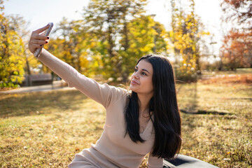 Naklejka premium Young woman taking selfie in autumn park using smartphone