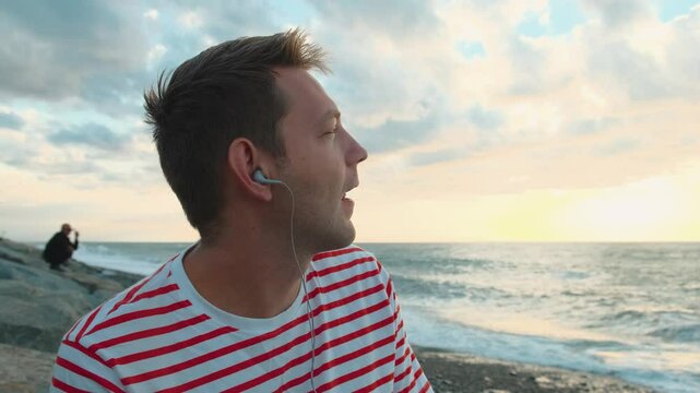 Happy Caucasian young man listening to musc with headphones using smartphone on the sea coast and enjoying sunset. Handsome male outdoors using mobile phone with earphones on the beach