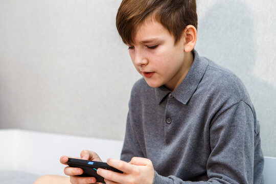 A ten year old boy is playing with his smartphone while sitting on the couch.