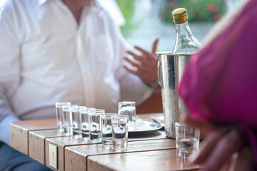 Close-up of a wooden table featuring shot glasses, a bottle in an ice bucket, and a blurred background suggesting an ambient restaurant environment during a gathering.