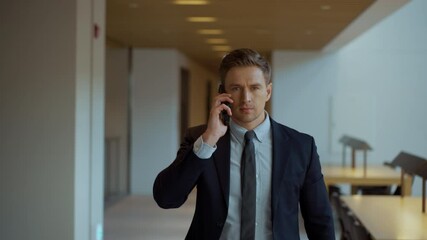 A serious businessman in his 30s picks up phone call while walking the office corridor - front view dolly pull back