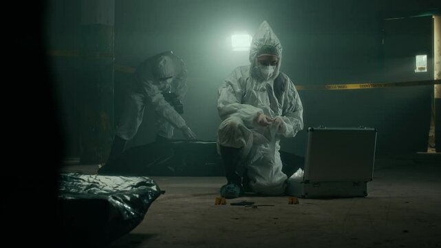 Full lowkey shot of two forensic experts wearing protective suits working in team at crime scene collecting evidence and examining corpse under black bag in dark deserted building