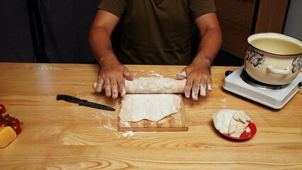Man preparing dinner. Adult man sitting at table at home in the kitchen and preparing spaghetti, rolls dough with rolling pin on the wooden table and board.