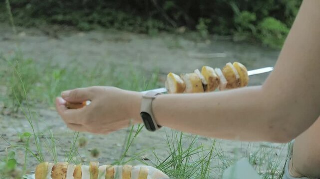 A woman strings pieces of potatoes and lard to fry over a fire.