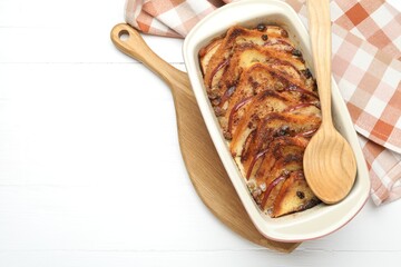 Delicious bread pudding in baking dish and spoon on white wooden table, top view. Space for text