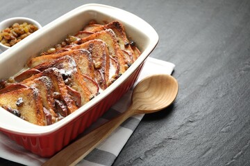 Freshly baked bread pudding in baking dish and wooden spoon on dark textured table, closeup. Space for text