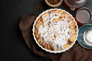 Delicious bread pudding with raisins, powdered sugar and tea on black table, flat lay