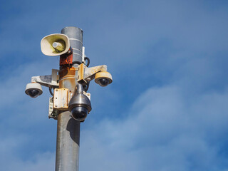 A pole with rusty, old CCTV cameras and loudspeaker on top of it. The sky is blue with some clouds in the background. Remote security and safety concept. Police monitoring point in a public place.