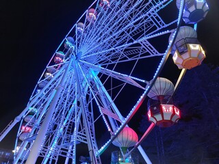 Ferris wheel dominates the frame, its white structure adorned with strings of green LED lights. The wheel is viewed from a low angle, emphasizing its height and the intricate network 