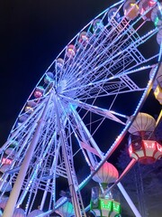 Ferris wheel dominates the frame, its white structure adorned with strings of green LED lights. The wheel is viewed from a low angle, emphasizing its height and the intricate network 