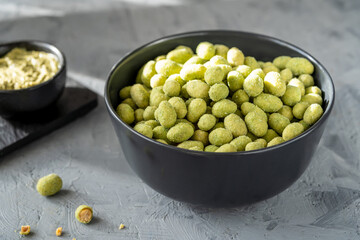 A black bowl filled with crunchy wasabi-coated peanuts, accompanied by a smaller bowl of dip, placed on a textured gray surface