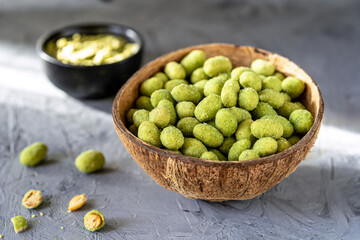 A rustic coconut bowl filled with wasabi-coated peanuts illuminated by soft natural light on a textured gray surface, with a black dish of wasabi dip in the background