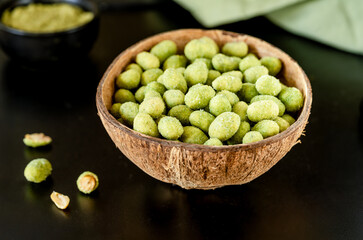 A coconut bowl filled with wasabi-coated peanuts on a sleek black surface, with scattered nuts and a green napkin in the background for a natural, stylish presentation