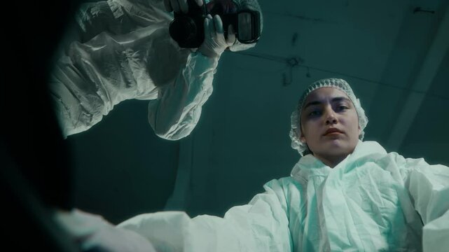 Bottom view shot of young Caucasian female forensic specialist in protective suit opening black body bag with African American male colleague taking photo on camera, discussing signs of struggle