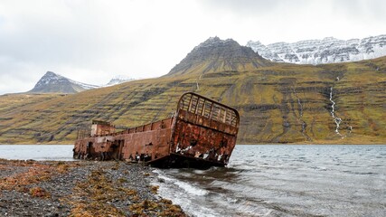 Rusted shipwreck on rocky shore with mountain backdrop.