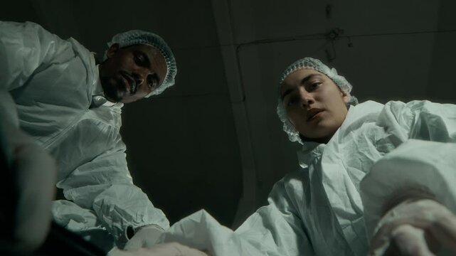 Bottom view lowkey shot of young diverse forensic colleagues wearing protective overalls looking down black body bag and discussing victim signs of struggle, working together at crime scene