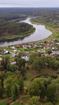 Aerial view capturing the winding Daugava river flowing gracefully through the residential area of Kraslava, Latvia, enveloped by lush green forests and a soft, cloudy sky, drone dolly in reveal shot