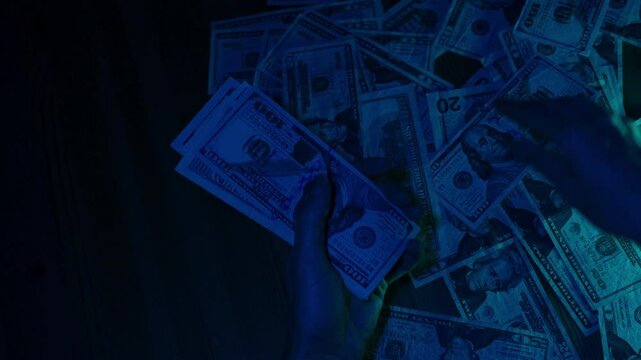 A woman hurriedly collects US dollar banknotes from a scattered pile on a table under dramatic blue lighting, conveying urgency and tension.