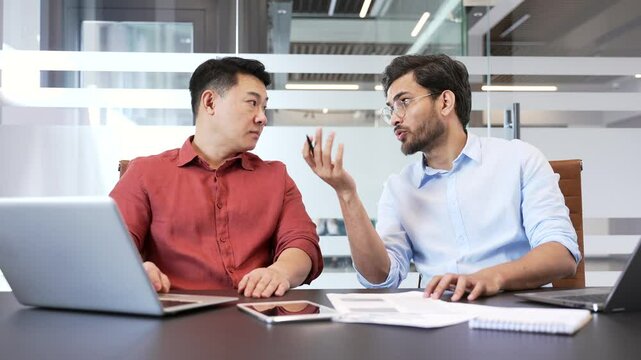 Two professional colleagues having a thoughtful discussion in modern office environment, with laptops and documents on the desk. Co-workers discuss work issues, talking about development project