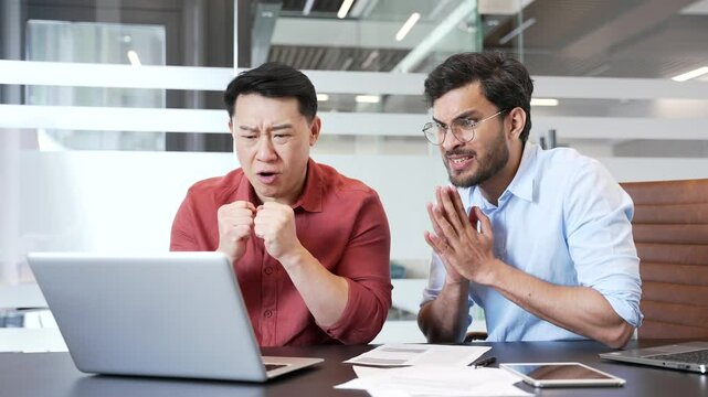 Two excited businessmen watching sports match or cheering for bids at auction using laptop while sitting in business office. Happy colleagues celebrating success showing excitement and achievement