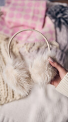 Furry earmuffs held in hand with soft knitted fabrics in background, emphasizing winter creativity and holiday spirit through warm accessories. Concept of cozy winter, style, aesthetic, relax.
