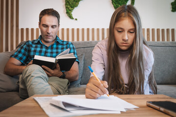Obraz premium A single father with a book in his hands helps his daughter to do her homework, girl writes the text. concept of parenting and education of children in a single-parent family, incomplete family