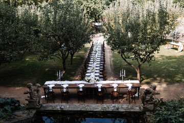 Long banquet table set for an outdoor event in a garden surrounded by apple trees. Elegant dining setup with white tableware, floral arrangements, and candelabras