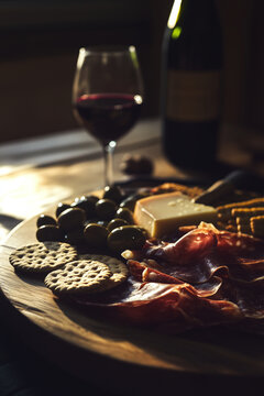 Wooden platter with cheeses, succulent slices of prosciutto and olives with glass of rich red wine in the background intimate vibe. Dark wood moody atmosphere