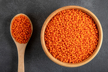 Red lentil grains in a wooden bowl and spoon on a gray background. Top view of dried orange lentils inside the bowl.